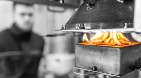 Cone calorimeter in operation: specimen burning with visible flames in the sample holder while the conical radiant heater supplies heat flux; a test operator is blurred in the background.