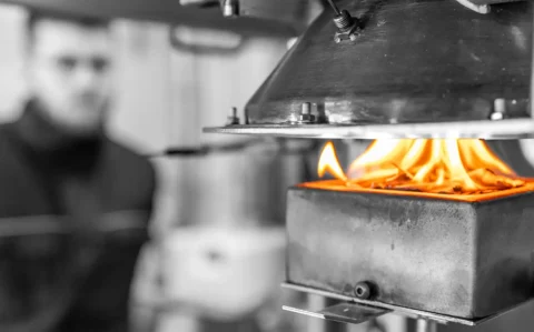 Cone-Kalorimeter im Einsatz: Eine Probe brennt mit sichtbaren Flammen im Probenhalter, darüber der konische Strahlungsheizer als Wärmeflussquelle; im Hintergrund unscharf ein Prüftechniker.