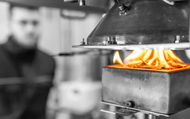 Cone-Kalorimeter im Einsatz: Eine Probe brennt mit sichtbaren Flammen im Probenhalter, darüber der konische Strahlungsheizer als Wärmeflussquelle; im Hintergrund unscharf ein Prüftechniker.
