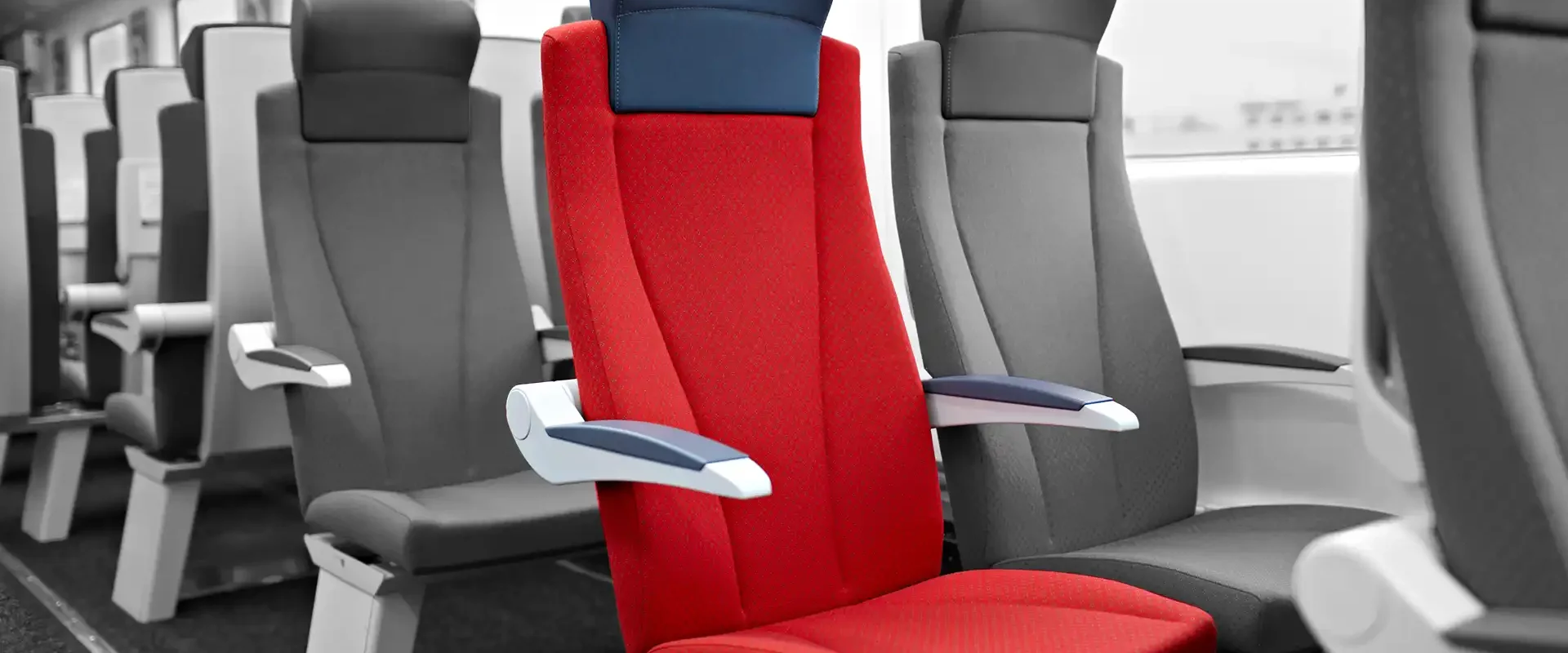 Roter Polstersitz mit blauer Kopfstütze in einer Zugkabine zwischen grauen Sitzen.