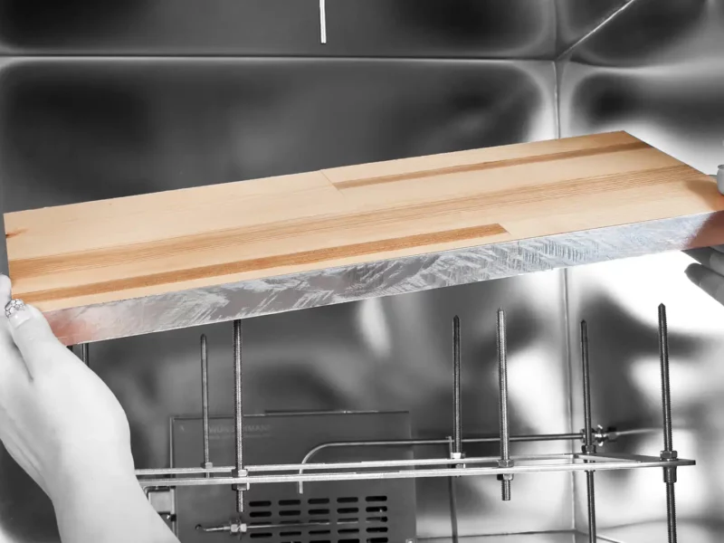 Zwei Hände setzen eine Holzplatte mit versiegelten Kanten in eine Edelstahl Emissionsprüfkammer.