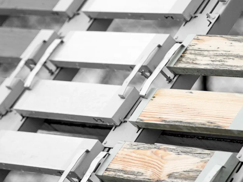 Metallgestell mit schräg montierten Holzplatten, teils mit Schimmel und Algen bewachsen, im Außenbewitterungsversuch.