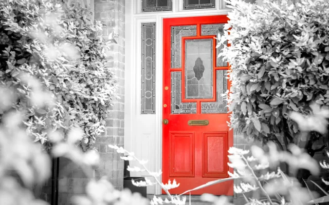 Rote Haustür mit verglasten Zierfeldern in einer Hausfassade, von Büschen eingerahmt.