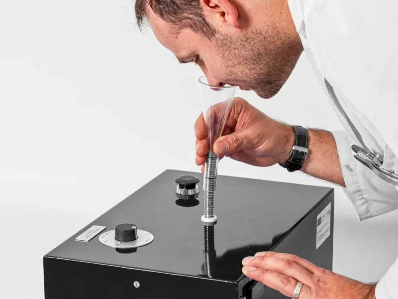 Laborant riecht an einem Trichteraufsatz eines Prüfgeräts zur Geruchsbestimmung von flüchtigen organischen Verbindungen