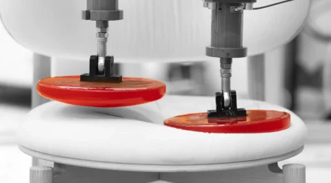 Two test actuators with red loading pads press on a padded chair seat—cyclic durability/fatigue test to assess indentation and long-term performance of furniture or automotive seats.