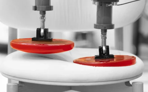 Zwei Prüfzylinder mit roten Lasttellern drücken auf die gepolsterte Sitzfläche eines Holzstuhls – Dauerbelastungs-/Ermüdungsprüfung zur Beurteilung von Verformung und Haltbarkeit von Möbel- bzw. Fahrzeugsitzen.