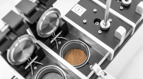 Laboratory tester with several circular measuring cells and closable lids; one cell contains a round wood specimen.