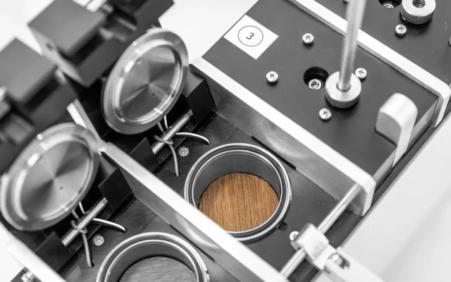 Labor-Prüfgerät mit mehreren runden Messkammern und verschließbaren Deckeln; in einer Kammer liegt eine runde Holzprobe