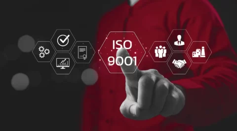 Hand pointing at a glowing virtual hexagon labeled “ISO 9001,” surrounded by icons for quality, processes, certificate, analytics, people, leadership, partnership and manufacturing