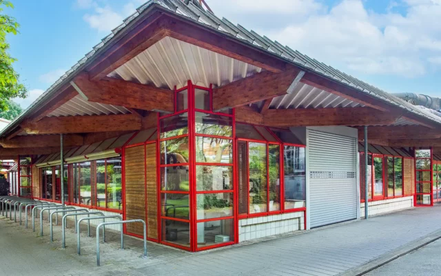 Außenaufnahme eines eingeschossigen Labor und Bürogebäudes mit roten Fensterprofilen, Holzfassade, weit auskragendem Dach und Fahrradständern.