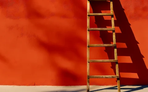 Holzleiter lehnt an einer orangefarbenen Wand und wirft einen deutlichen Schatten.